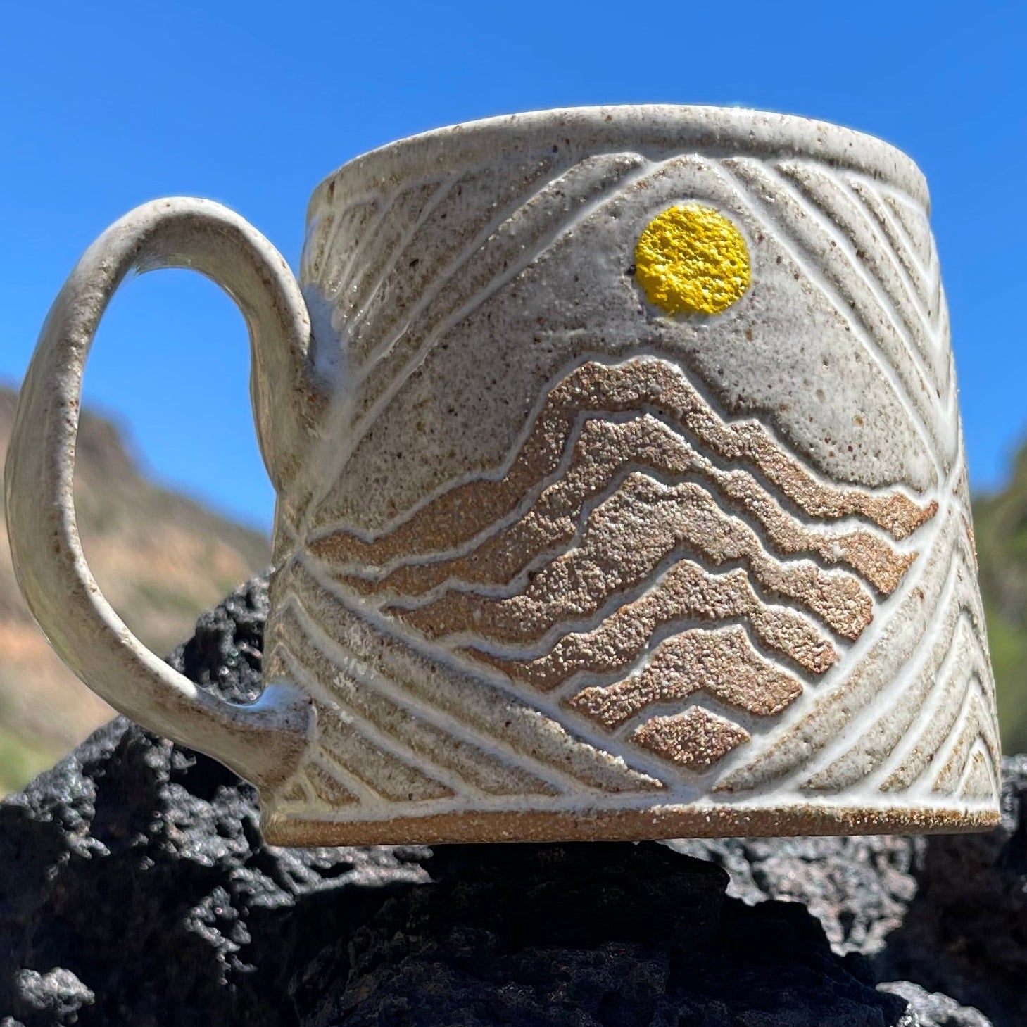 A handmade ceramic mug with a textured beige finish, featuring a hand-painted tan wave pattern and a small yellow sun accent, resting on a dark volcanic rock with a mountain and blue sky in the background.